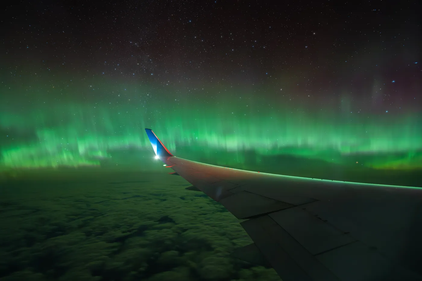 Aurora seen from an aircraft window, above the cloud, with the wing light lit.