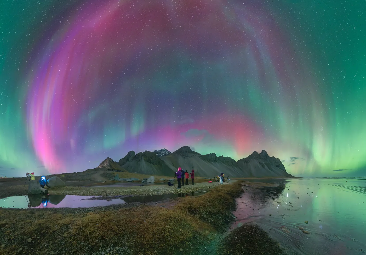 A pink and violet corona over jagged peaks, with small figures and their reflection.