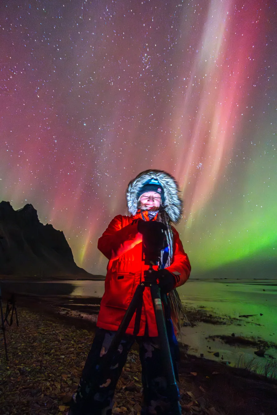 Cari Letelier in a red parka behind her tripod, under red and green aurora on the coast.