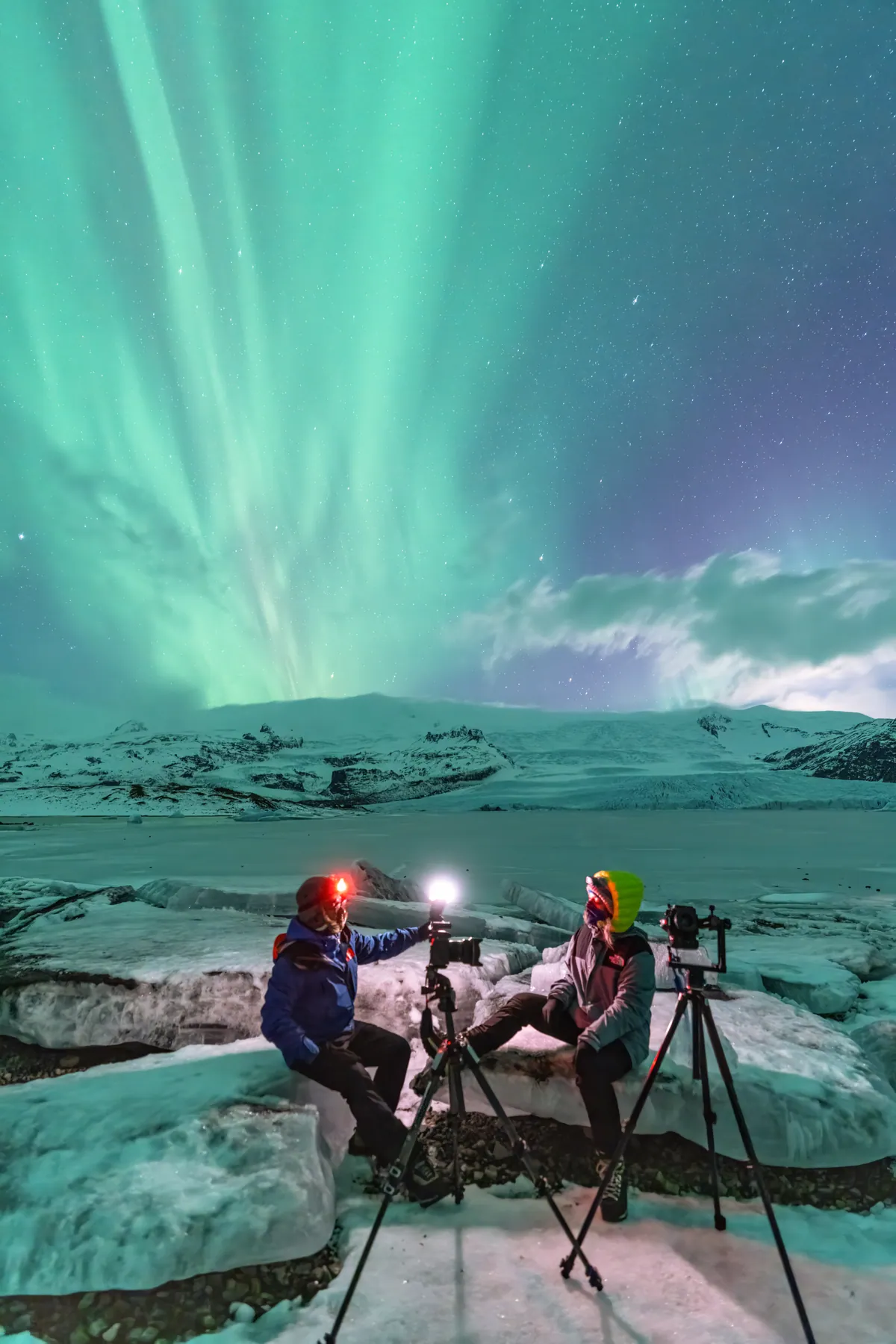 Cari and Mario with headlamps on, working with two tripods on glacier ice under a green aurora.