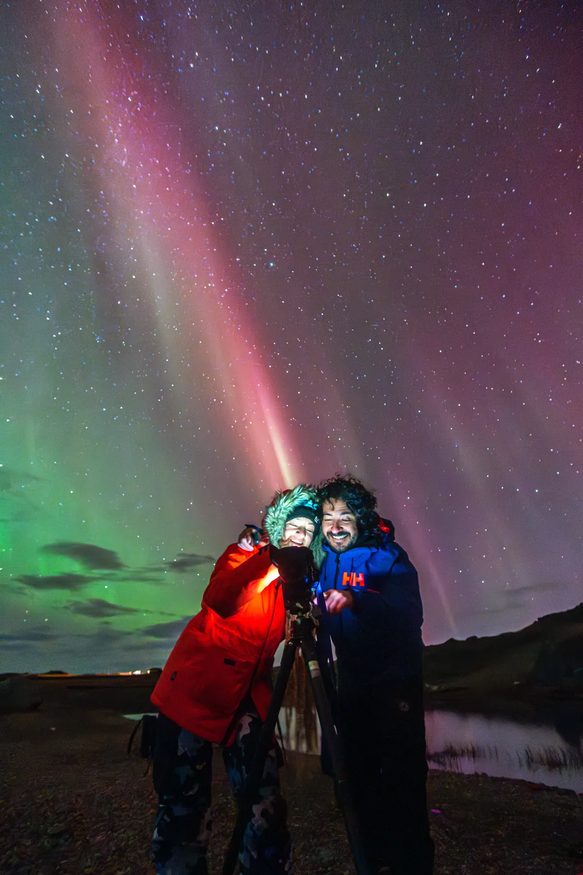 The two of them laughing behind a camera under red and green aurora rays.