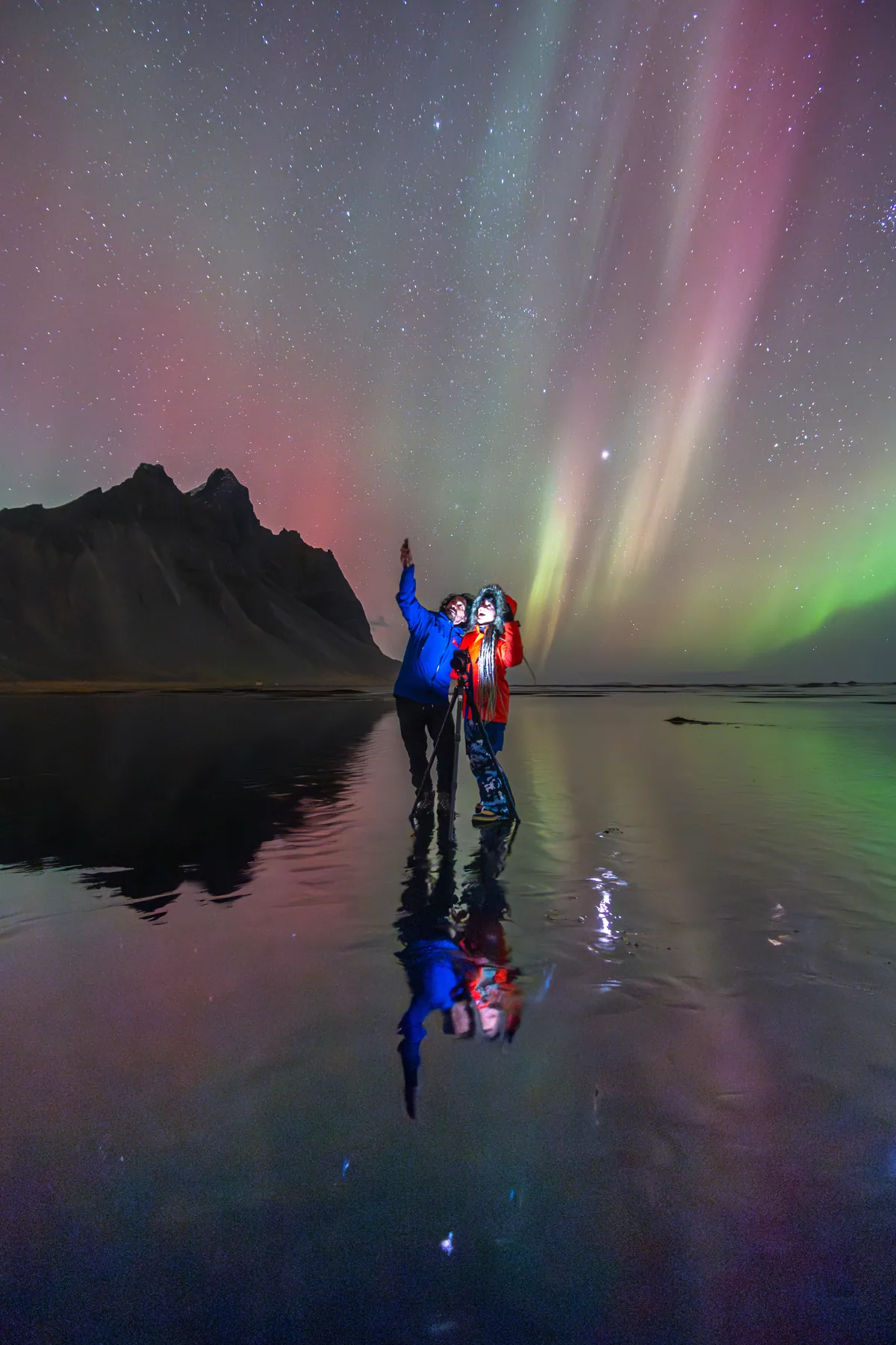 Two tiny figures beside a tripod under a pink and green aurora mirrored in still water.