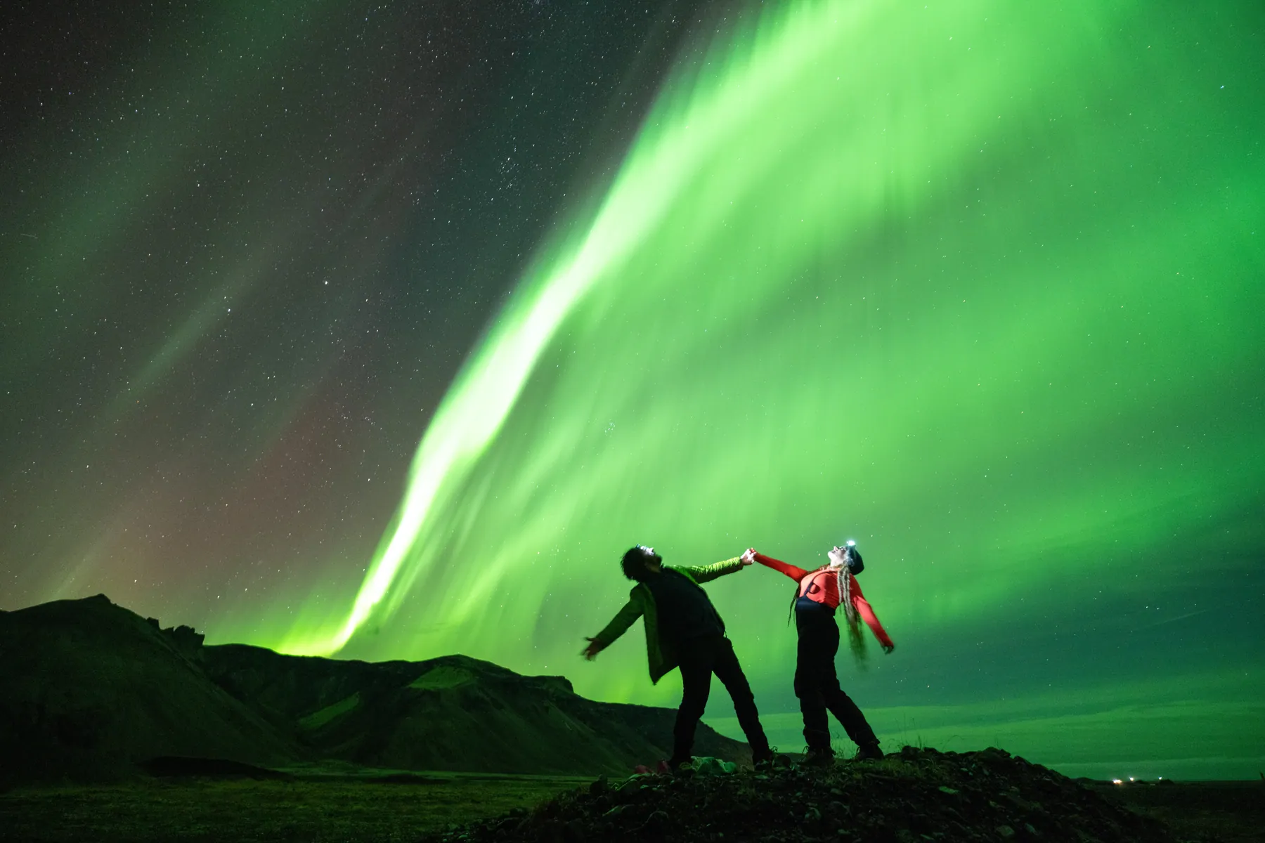 Two silhouettes holding hands on a ridge under a wide green band of aurora.