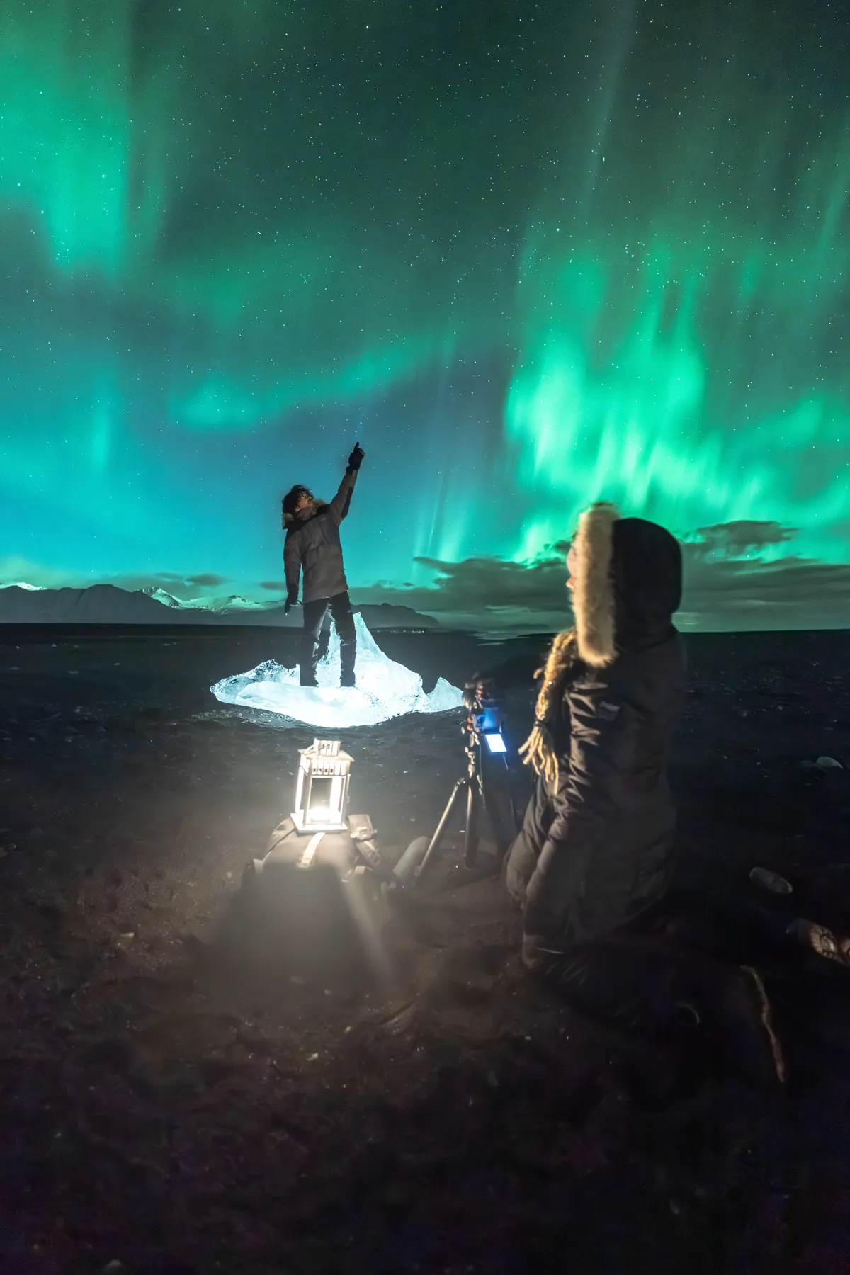 One of them pointing at the sky beside a tripod under red and green aurora.