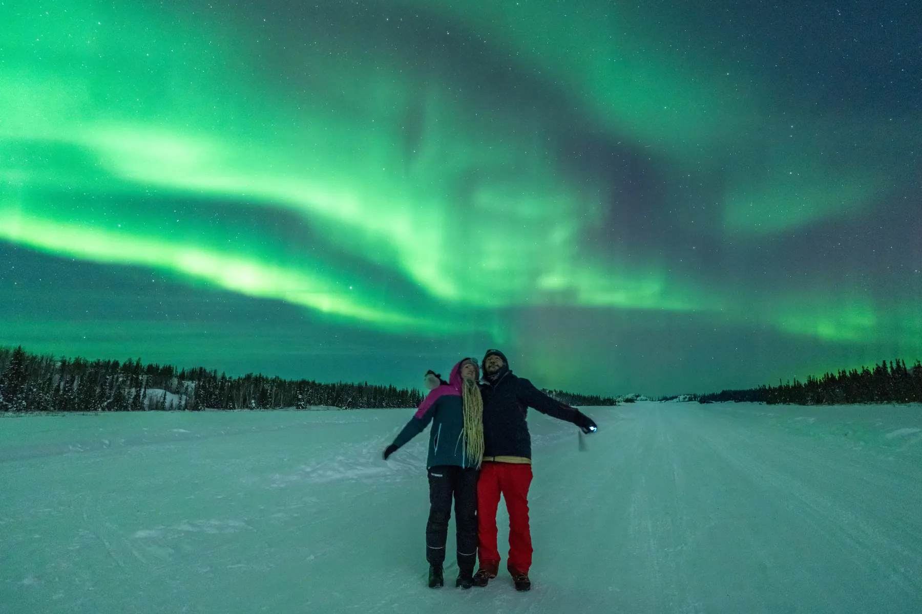 The two of them with arms open on a snowy road under bands of green aurora.