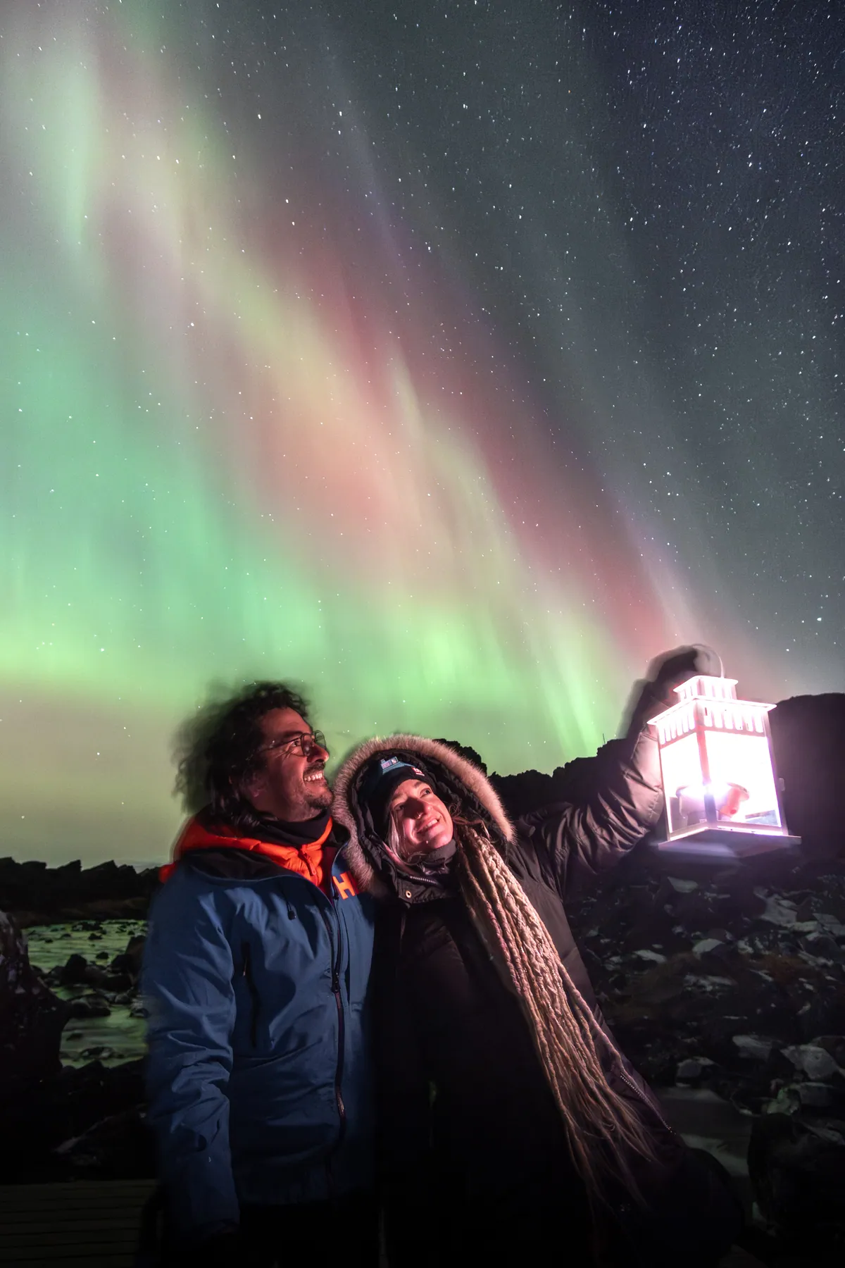 The two of them beside a lit panel under green and pink aurora by the sea.