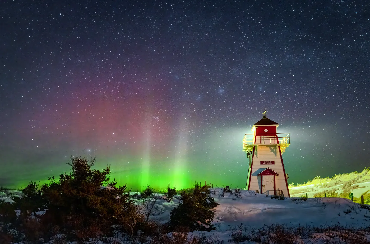 A red and white lighthouse in the snow with green aurora low on the horizon.