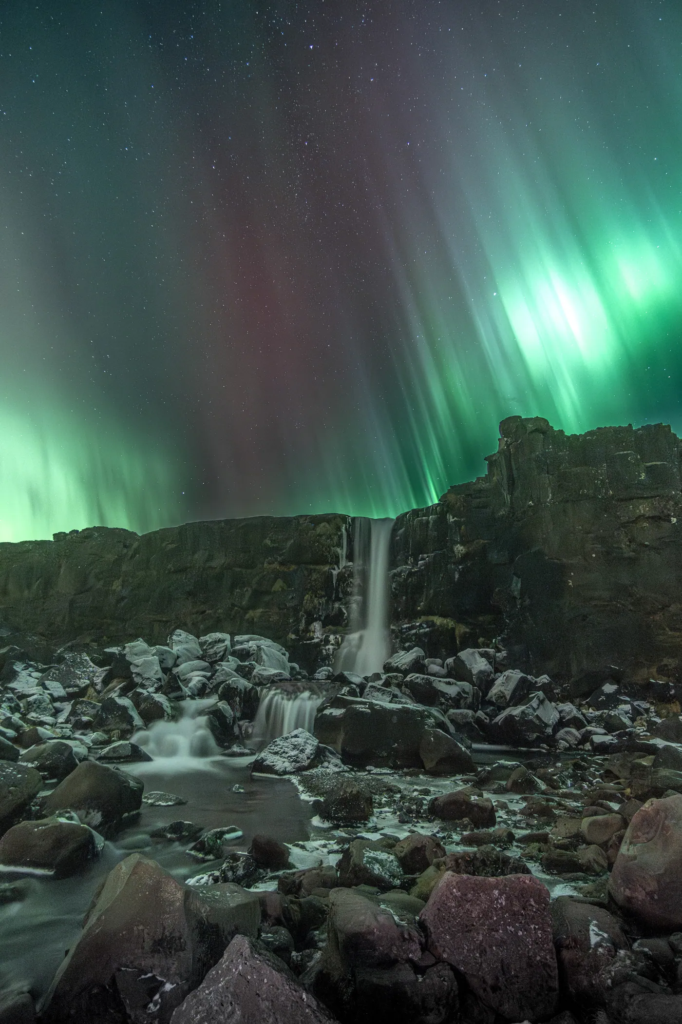 A waterfall between rocks under green and red aurora rays.